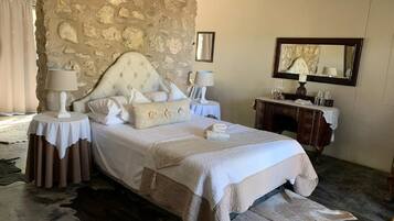 Une chambre avec un mur en pierre, un lit recouvert de draps blancs, deux tables de chevet avec des lampes, une commode en bois et un miroir.