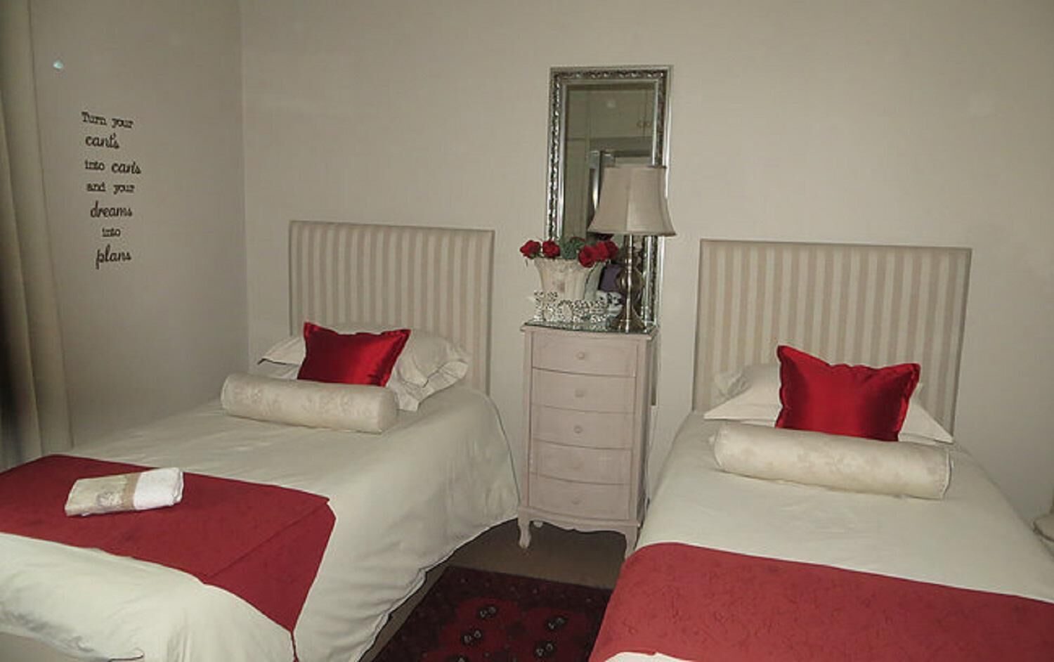 Une chambre d’hôtel avec deux lits, chacun agrémenté d’un oreiller rouge et d’une couverture blanche. On y trouve une commode surmontée d’un miroir, ainsi qu’un mur orné d’une citation inspirante.