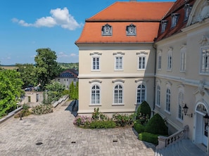 A building with a red-tiled roof, multiple windows, and a paved courtyard with a small garden. - La Contessa Castle Hotel (Szilvasvarad)