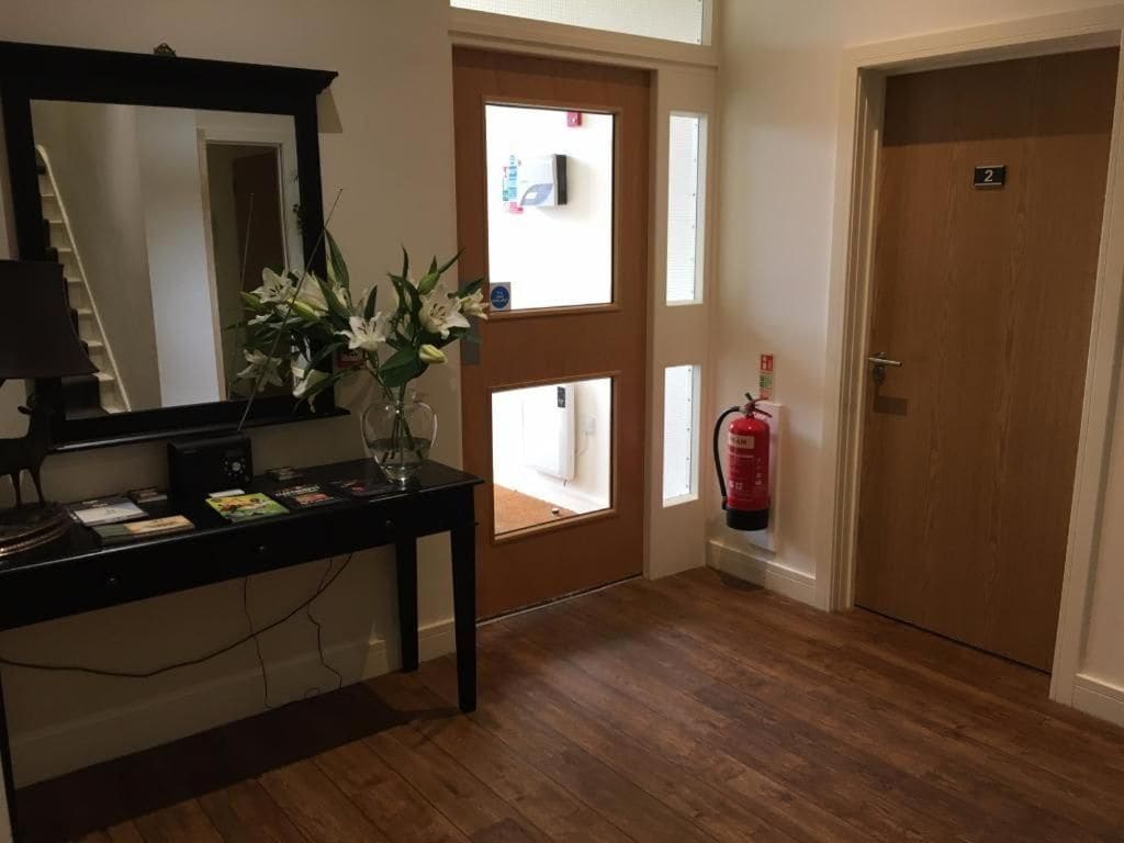Habitación de hotel con piso de madera, un escritorio con lámpara, un espejo, un florero con flores, un extintor y una puerta con un letrero con número.