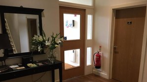 A hotel room with a wooden floor, a desk with a lamp, a mirror, a vase with flowers, a fire extinguisher, and a door with a number sign. - The Burlington Hotel (Cleethorpes)