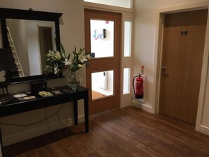 A hotel room with a wooden floor, a desk with a lamp, a mirror, a vase with flowers, a fire extinguisher, and a door with a number sign.