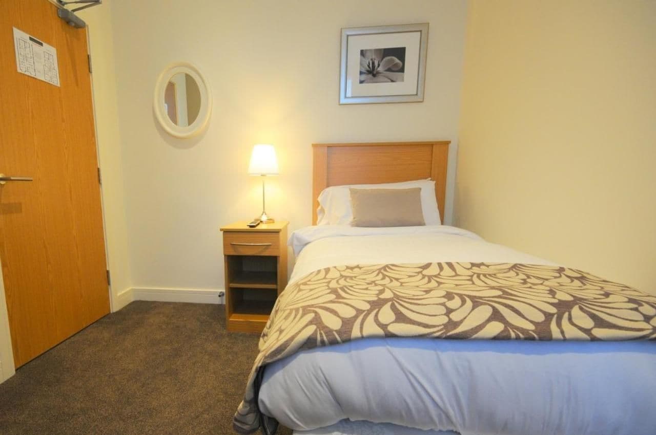 Une chambre d’hôtel avec un lit, une lampe de chevet, une petite table de nuit en bois, un miroir et un tableau encadré au mur.