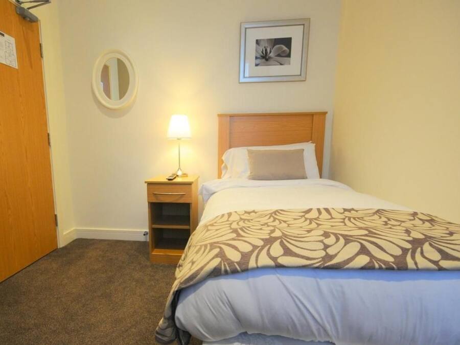 Une chambre d’hôtel avec un lit, une lampe de chevet, une petite table de nuit en bois, un miroir et un tableau encadré au mur.