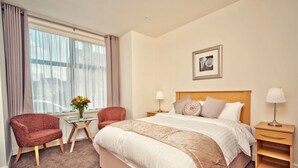 A hotel room with a large bed, two armchairs, a small table with a vase of flowers, a bedside table with a lamp, and a framed picture on the wall. - The Burlington Hotel (Cleethorpes)