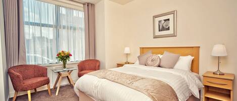 Una habitación de hotel con una cama grande, dos sillones, una mesita con un florero, una mesita de noche con una lámpara, y un cuadro en la pared.