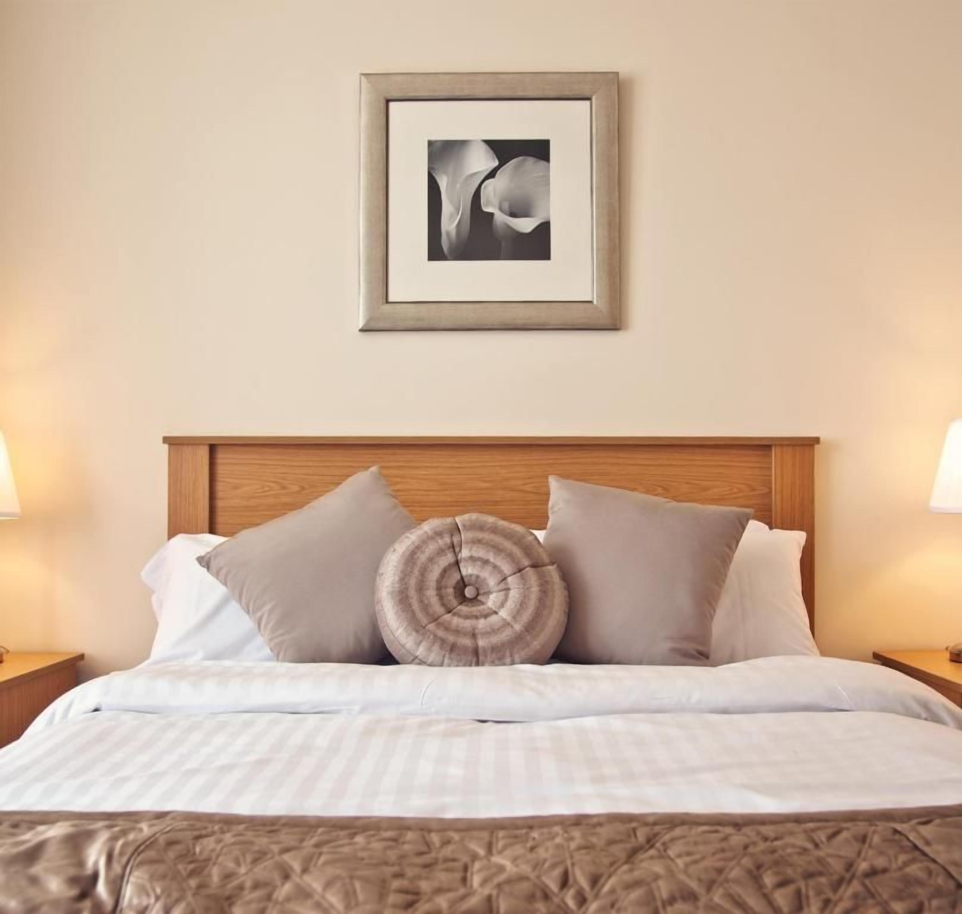 A neatly made bed with a wooden headboard, a framed picture above it, and two bedside lamps on either side.