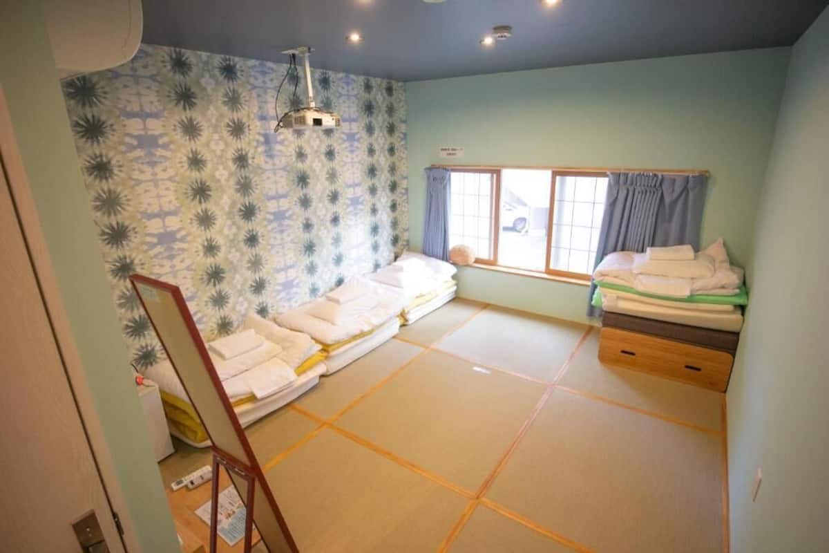 Una habitación con suelo de tatami, una cama con ropa de cama blanca, una ventana con cortinas y un proyector instalado en el techo.