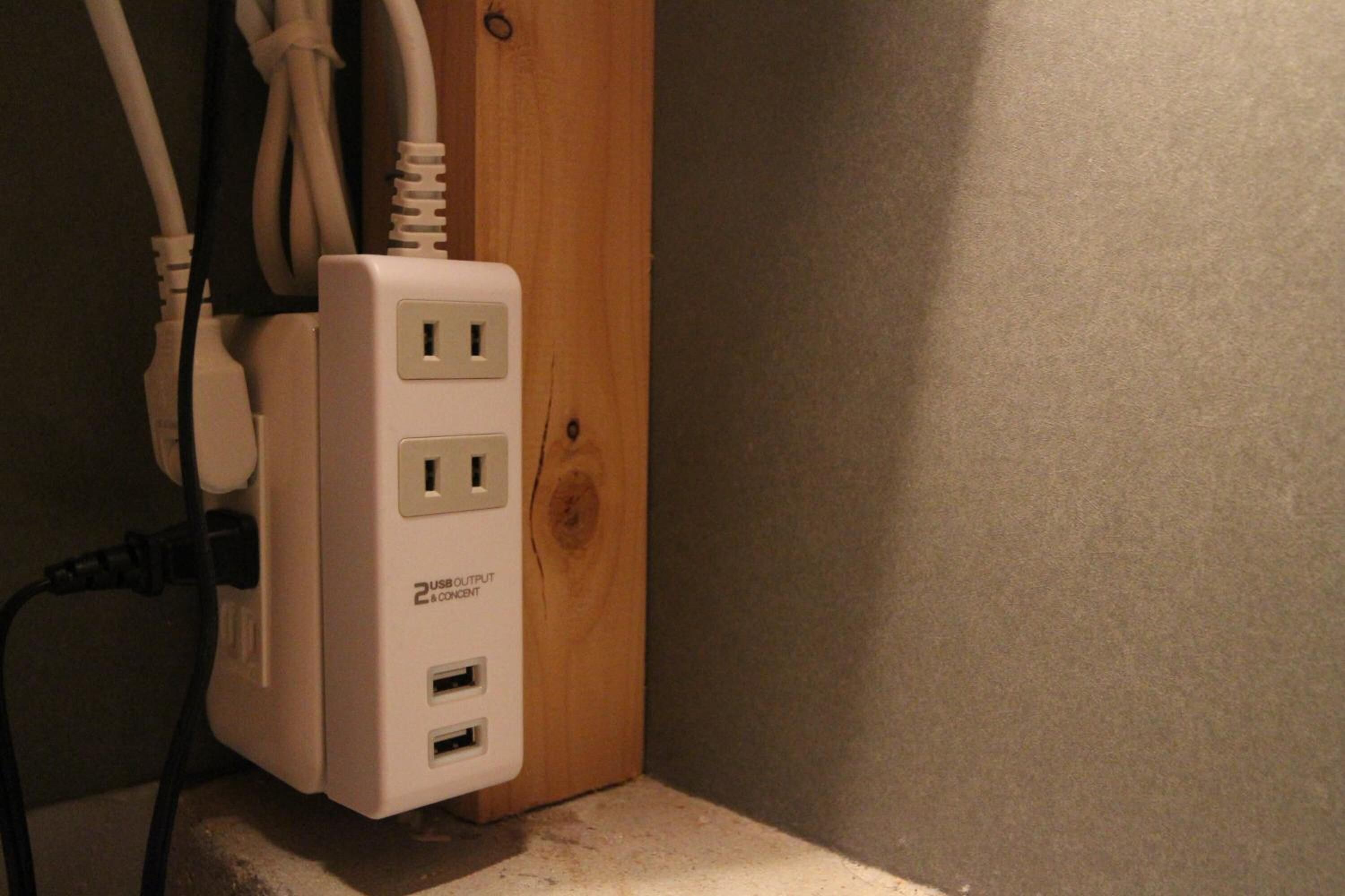 A white USB output power strip with two USB ports and two electrical outlets, mounted on a wooden surface against a wall.