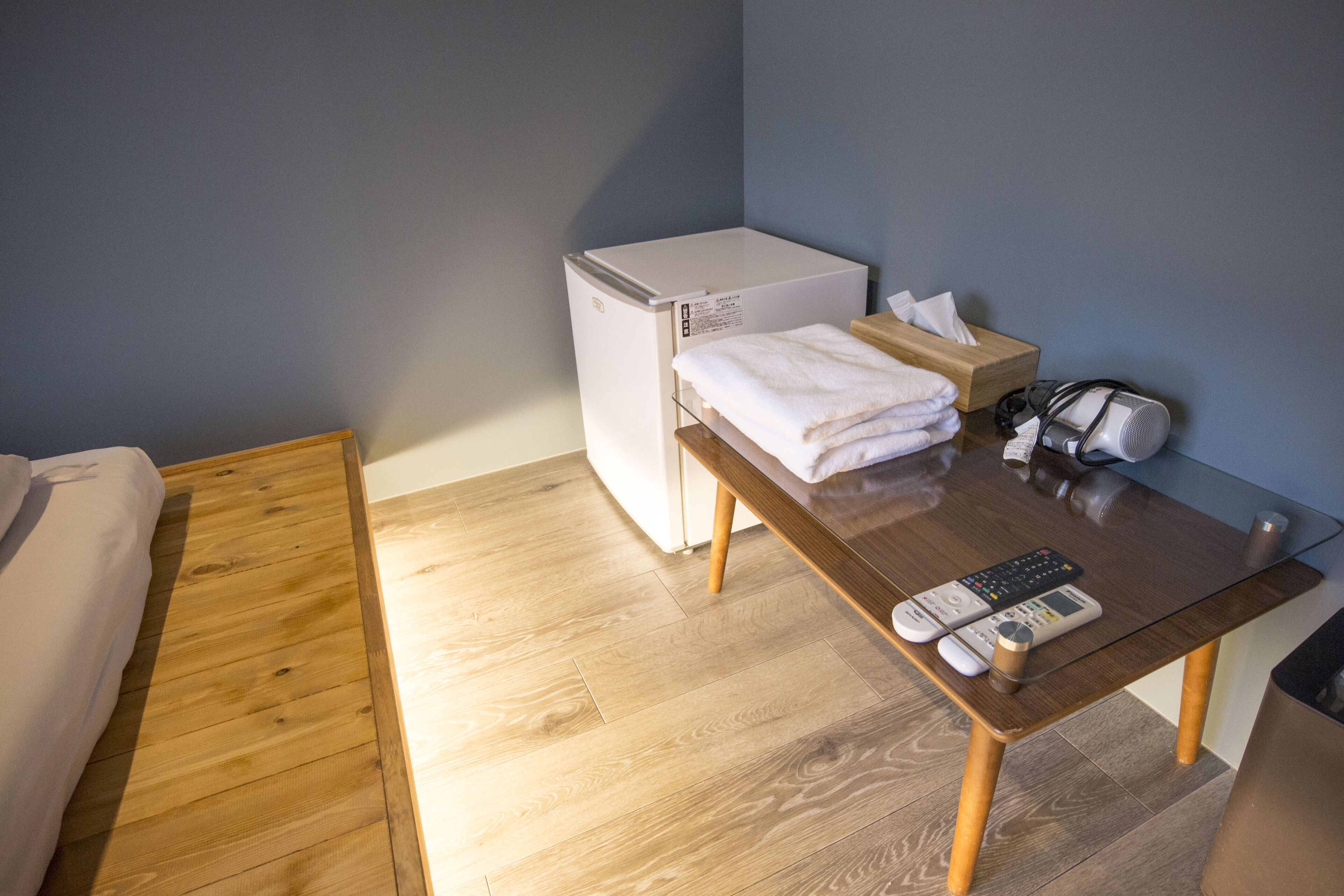 Una habitación compacta con una cama, un refrigerador blanco pequeño, una mesa de madera con un control remoto, una caja de pañuelos y un secador de pelo.