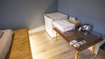 Una habitación compacta con una cama, un refrigerador blanco pequeño, una mesa de madera con un control remoto, una caja de pañuelos y un secador de pelo.