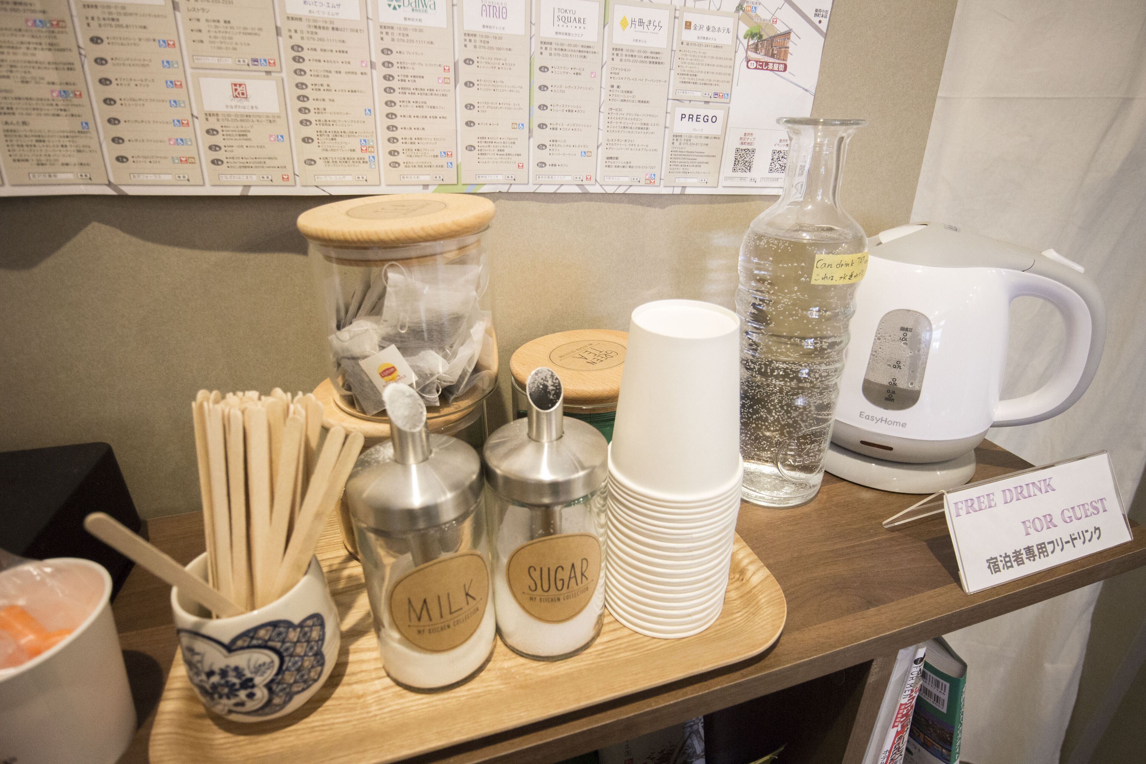Una barra de desayuno con utensilios, recipientes para leche y azúcar, un vaso de agua, una máquina de café y un cartel que ofrece una bebida gratuita a los huéspedes.