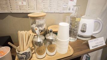 Una barra de desayuno con utensilios, recipientes para leche y azúcar, un vaso de agua, una máquina de café y un cartel que ofrece una bebida gratuita a los huéspedes.