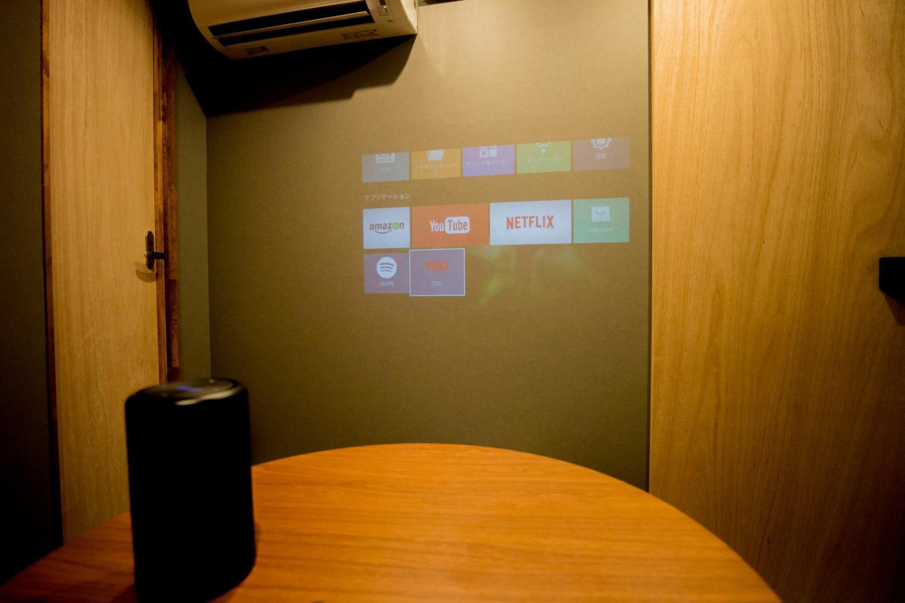 A room with a wooden table, a black speaker, and a wall projection displaying various streaming services.