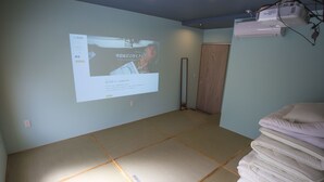 A room with tatami flooring, a projection screen displaying a webpage, and a stack of pillows. - SHARIN Kanazawa Traveler's Inn - Hostel (Kanazawa)