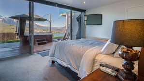 Une chambre moderne avec un grand lit, une table de chevet en bois, une télécommande et une lampe. Une porte-fenêtre coulissante donne accès à un espace extérieur aménagé avec un banc et une table.