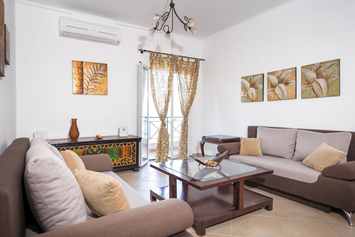 A living room with a sofa, a coffee table, and a TV. There is a window with curtains and a painting on the wall.