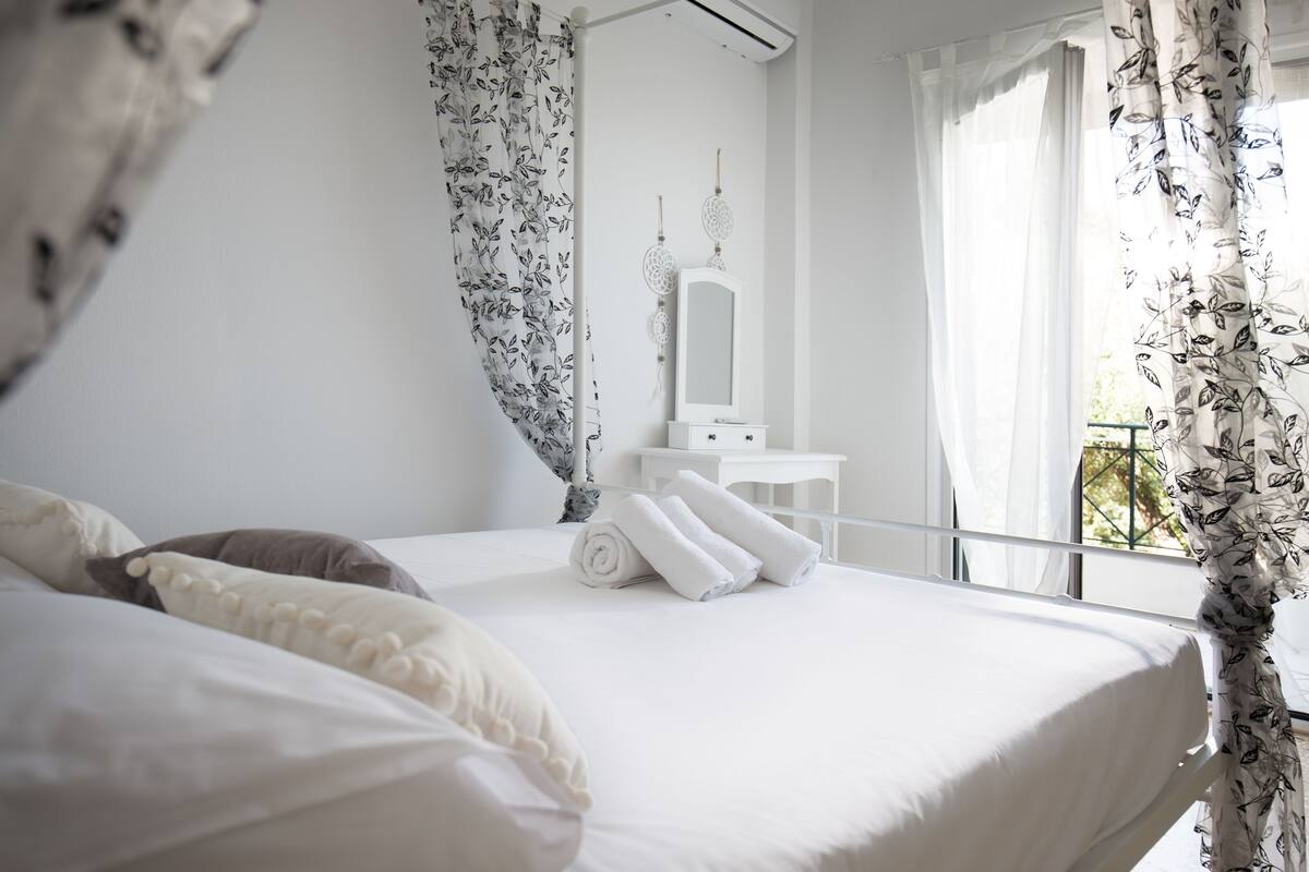 A neatly made bed with white linens and pillows, a white bedside table with a mirror and a small decorative item, and a window with floral curtains.
