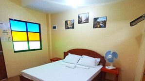 A hotel room with a wooden bed, a nightstand with a fan, and a window with a colorful view. - Pamujo Hostel (Baclayon)