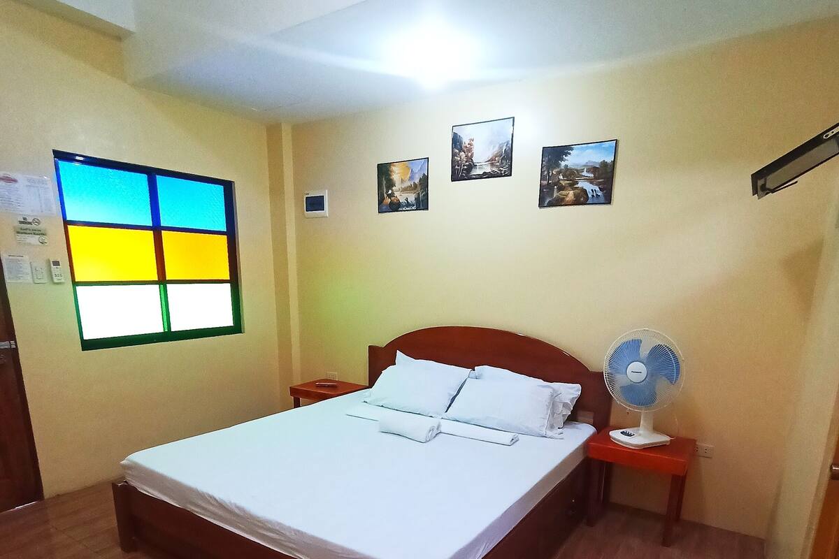 Habitación de hotel con cama de madera, una mesita de noche con un ventilador y ventana con vistas coloridas.