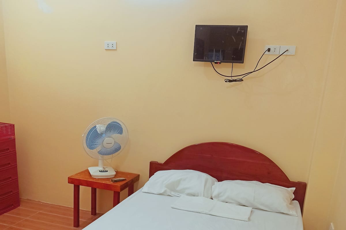 Un dormitorio con cama de madera, ropa de cama blanca, cabecera roja, televisor montado en la pared y ventilador de pie.
