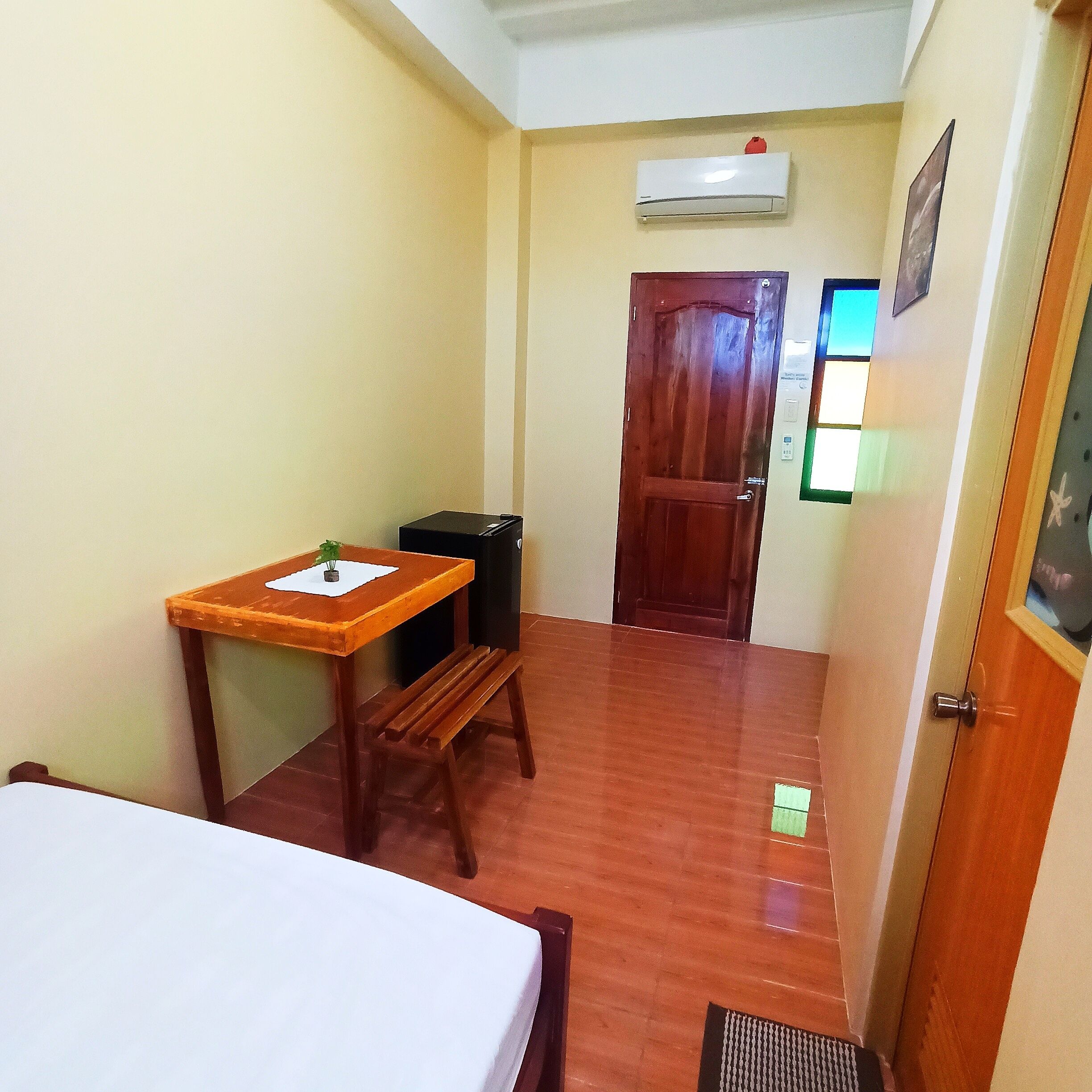 A small room with a bed, a wooden table with a plant, a bench, a door, and an air conditioning unit on the wall.