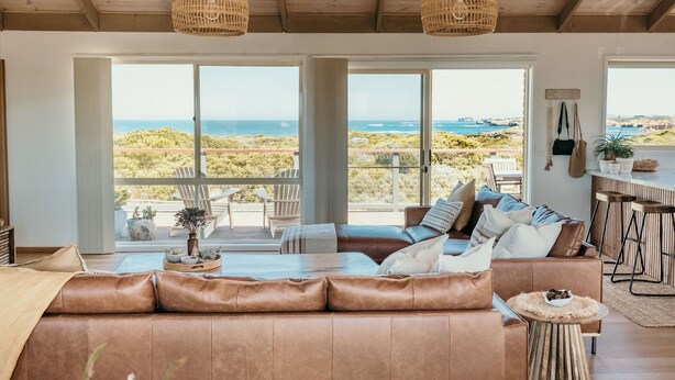 A spacious living area with a large brown leather sofa, two wicker pendant lights, and a view of the ocean through large windows.