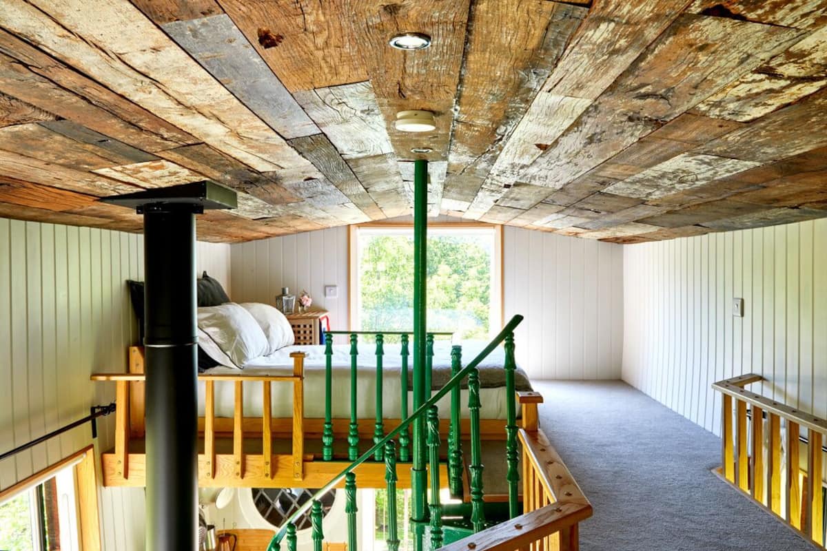A rustic room with a wooden ceiling, a staircase with green balusters, and a window providing natural light.