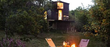 A rustic cabin with a fire pit and two lounge chairs in a wooded area.
