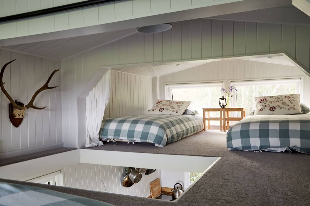 A loft bedroom with two beds, a wooden ceiling, and a kitchen area below.