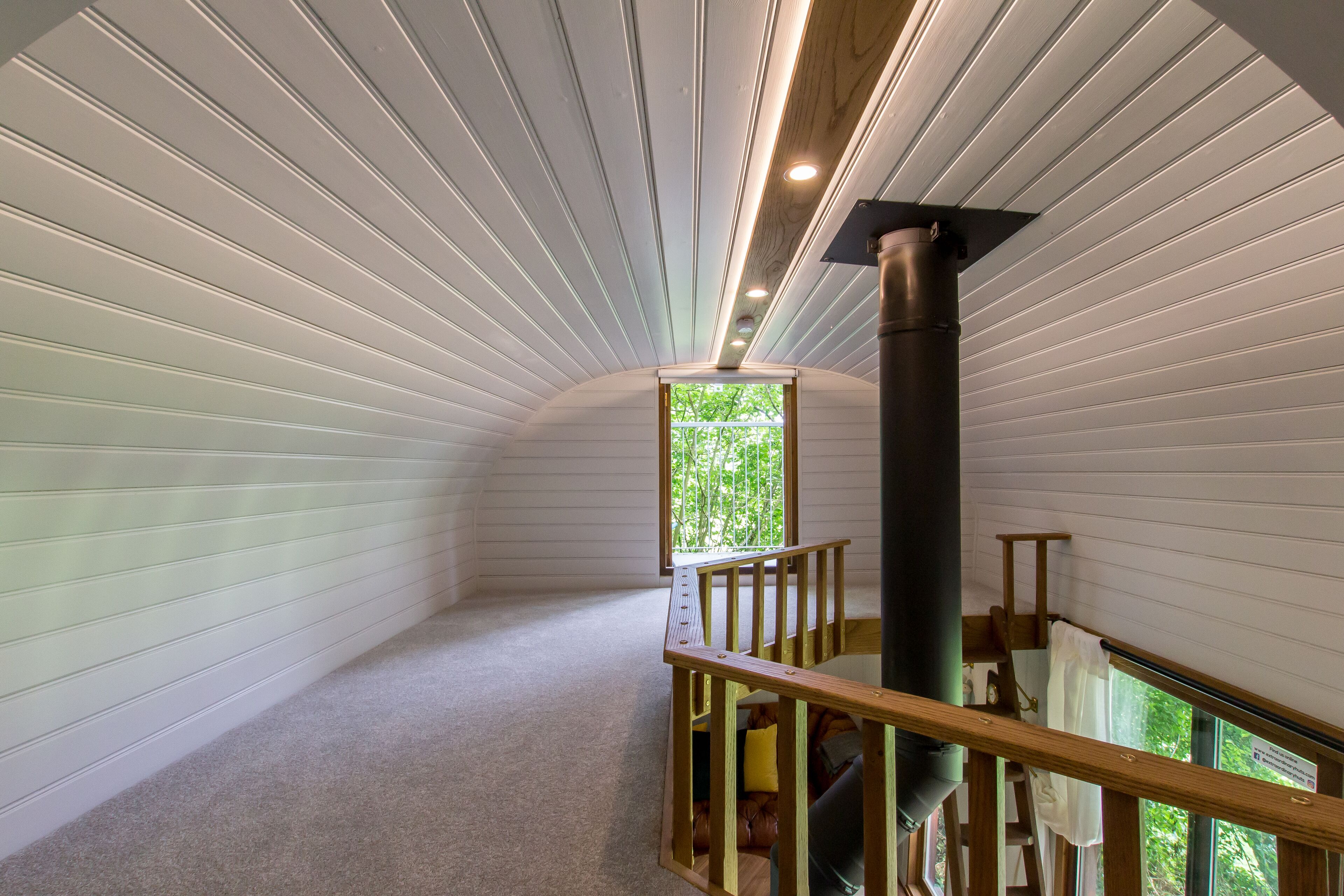 A room with white panel walls, a wooden staircase, and a window with a view of greenery.