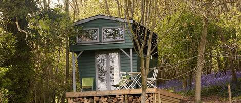 A small, green wooden cabin with a balcony, surrounded by trees and purple flowers.