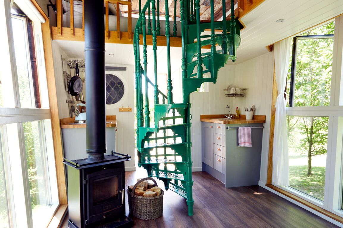 A cozy kitchen with a wood-burning stove, a green spiral staircase, and a large window offering a view of the outdoors.