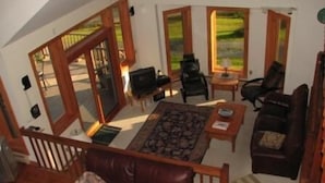A cozy living room with a brown leather sofa, a coffee table, and a TV. There are large windows with a view of greenery outside. - Franconia Notch Retreat, Huge Views, Hot Tub, EV charger, A/C (Sugar Hill)