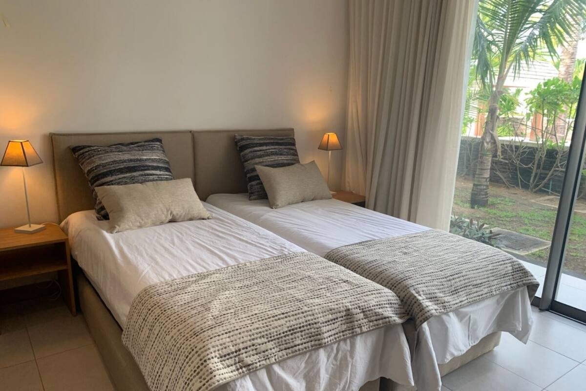 Ein Doppelbett mit zwei Kissen, ein Nachttisch mit Lampe und ein großes Fenster mit Vorhängen.