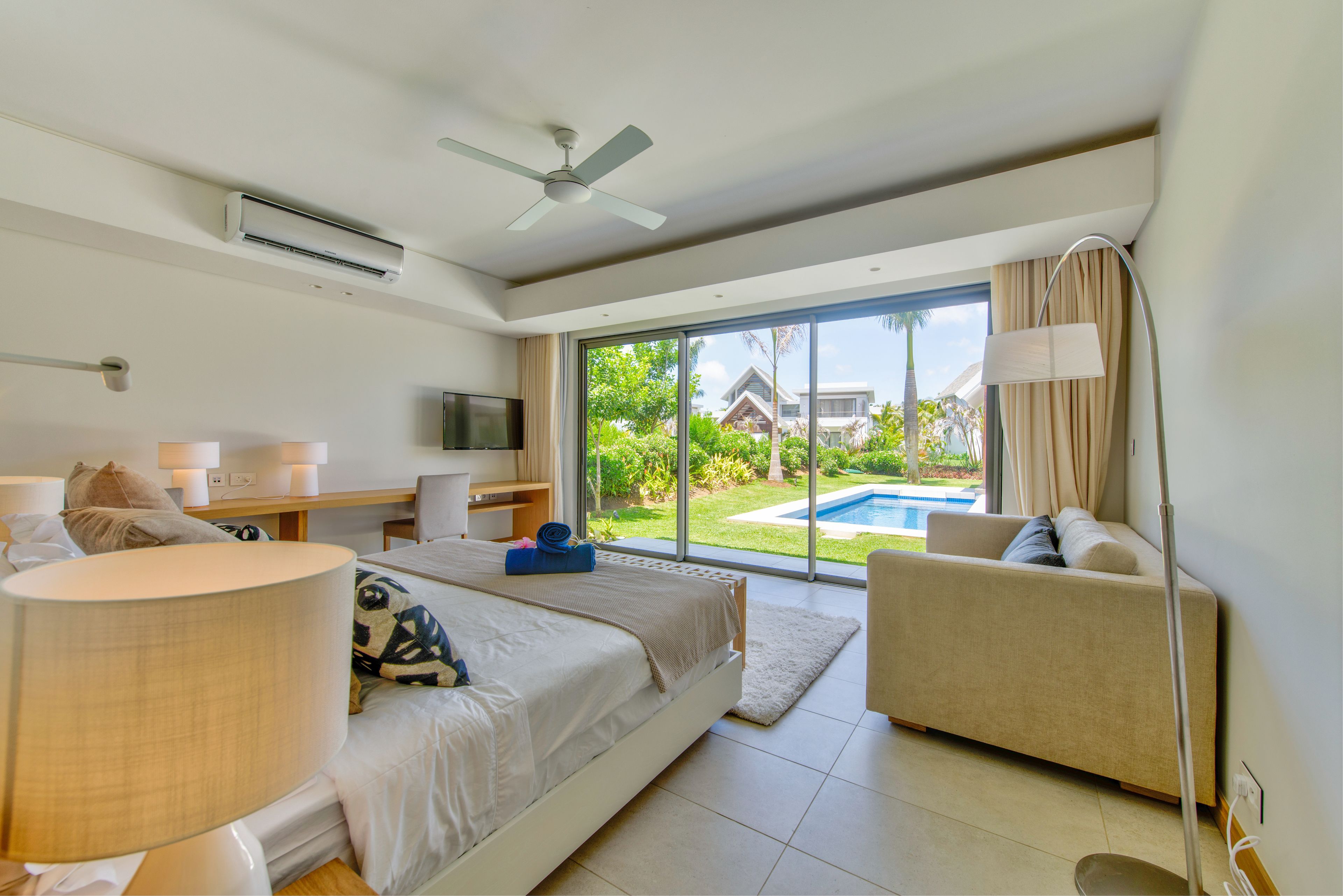 A modern hotel room with a bed, a sofa, a desk, and a large window offering a view of a swimming pool and greenery.
