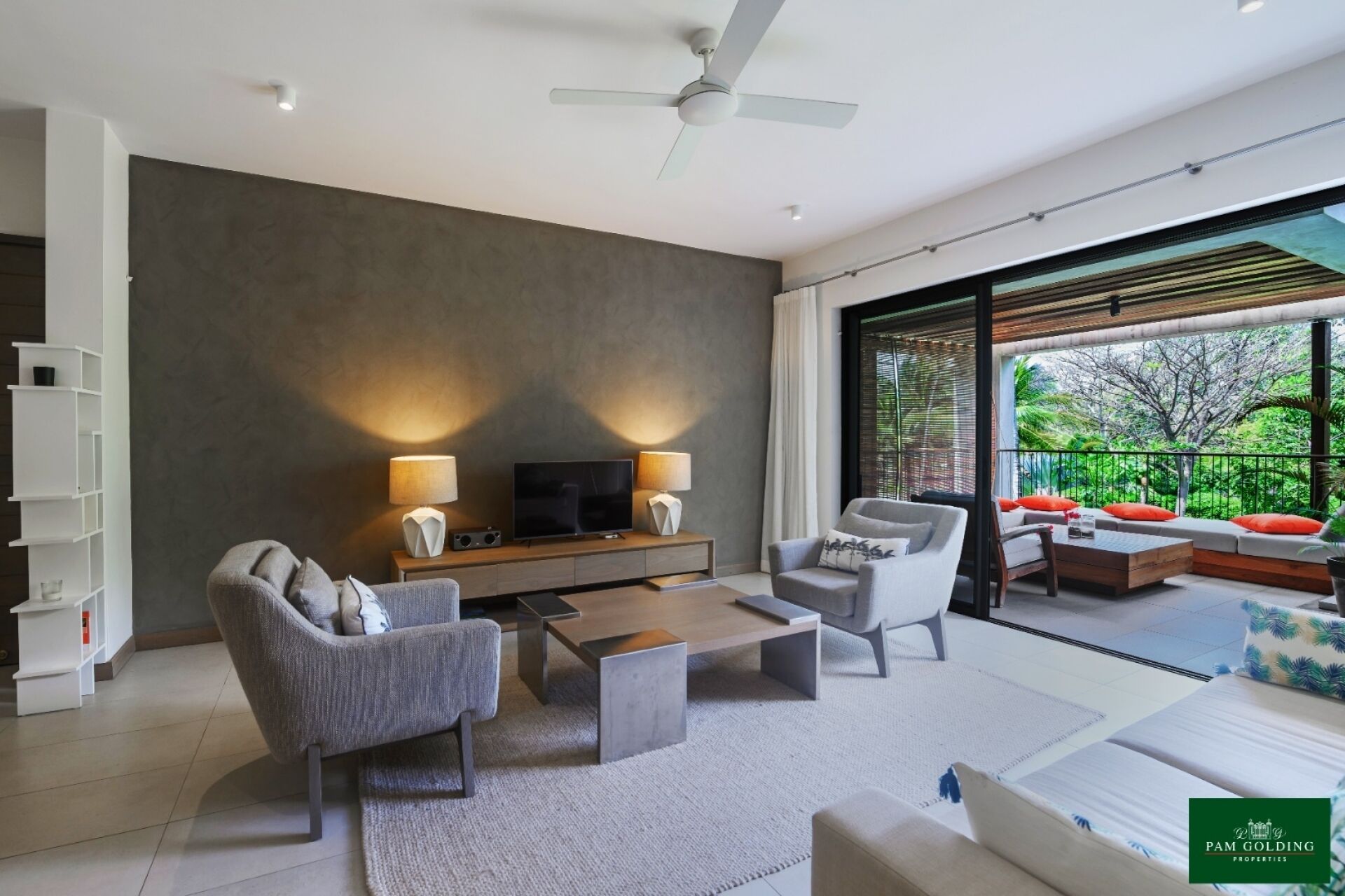 A modern living room with a flat-screen TV, two lamps, a coffee table, and a sofa. There is a bookshelf and a large window with a view of greenery.
