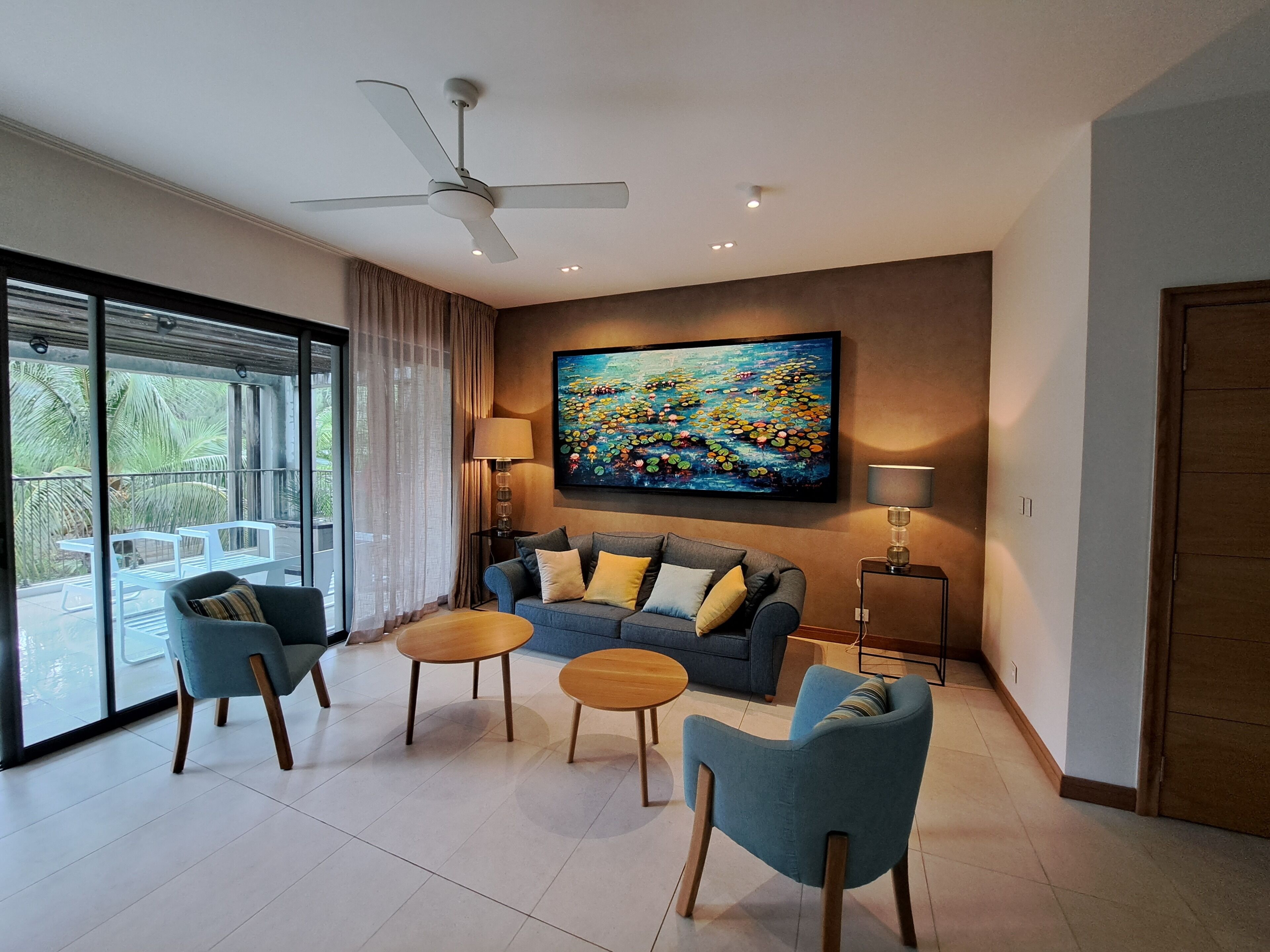 A modern living room with a sofa, two armchairs, a coffee table, a ceiling fan, a painting, and a view through large windows.