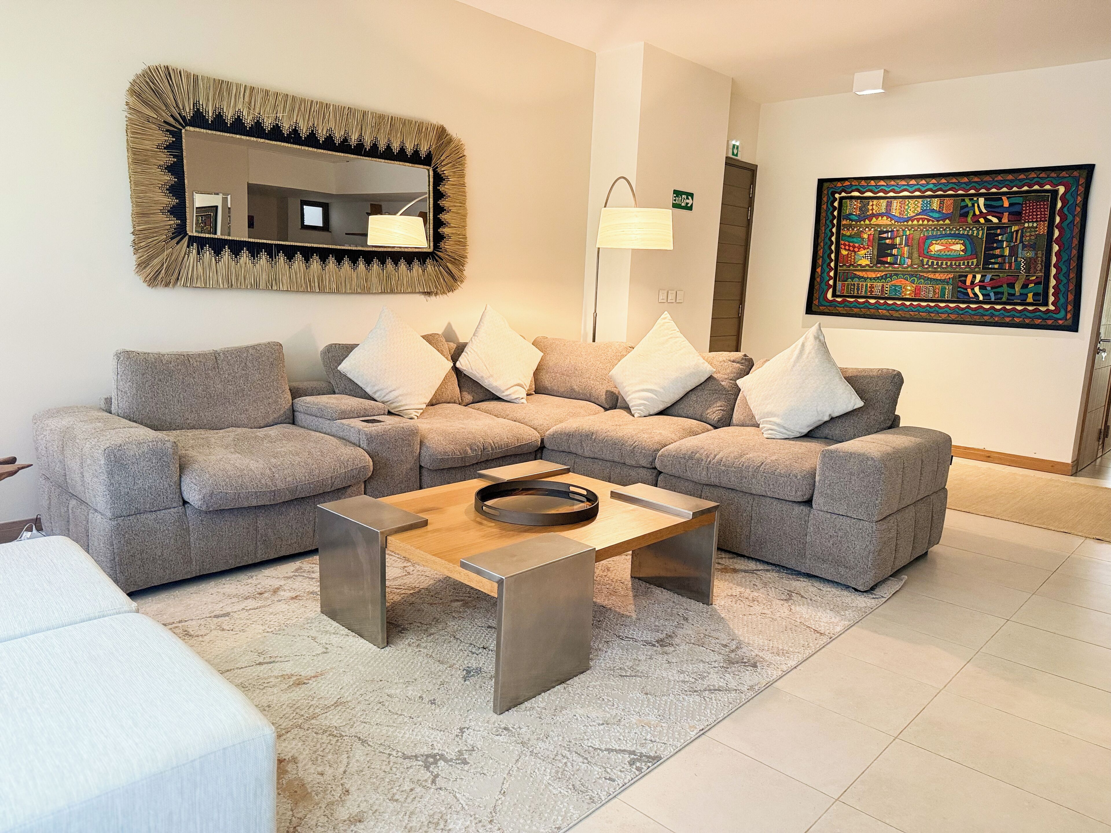 Una sala de estar moderna con un sofá modular, una mesa de centro, un espejo montado en la pared, una lámpara de pie y una obra de arte colorida.