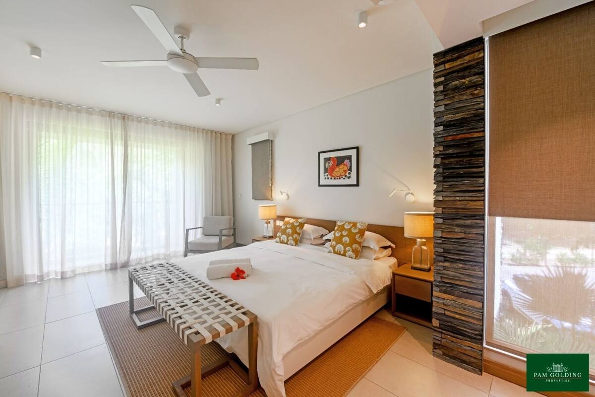 Ein Hotelzimmer mit Bett, Sessel, Schreibtisch, Deckenventilator, einem Fenster mit Vorhängen, einem Wandbild und einer Wandverkleidung aus Stein.