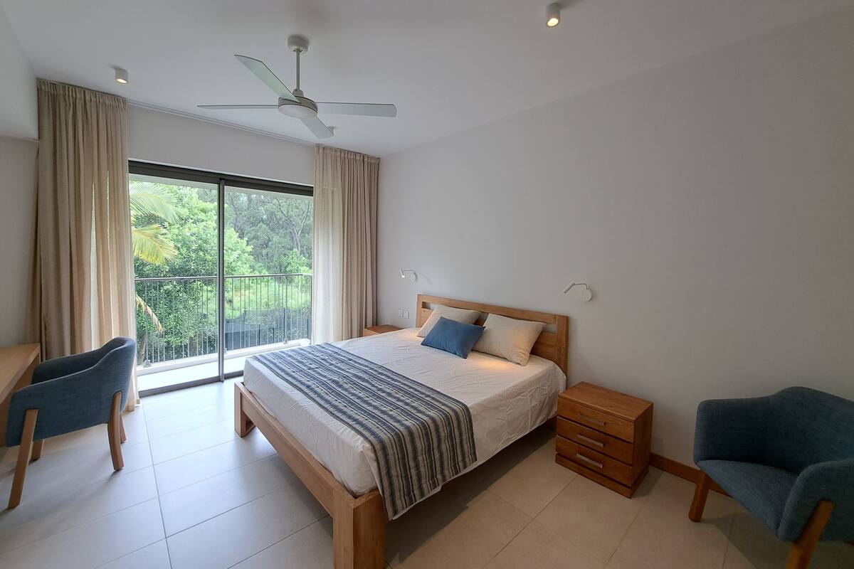 Ein Schlafzimmer mit Bett, Schreibtisch, Stuhl, Nachttisch und einem Balkon mit Blick auf Grünflächen.