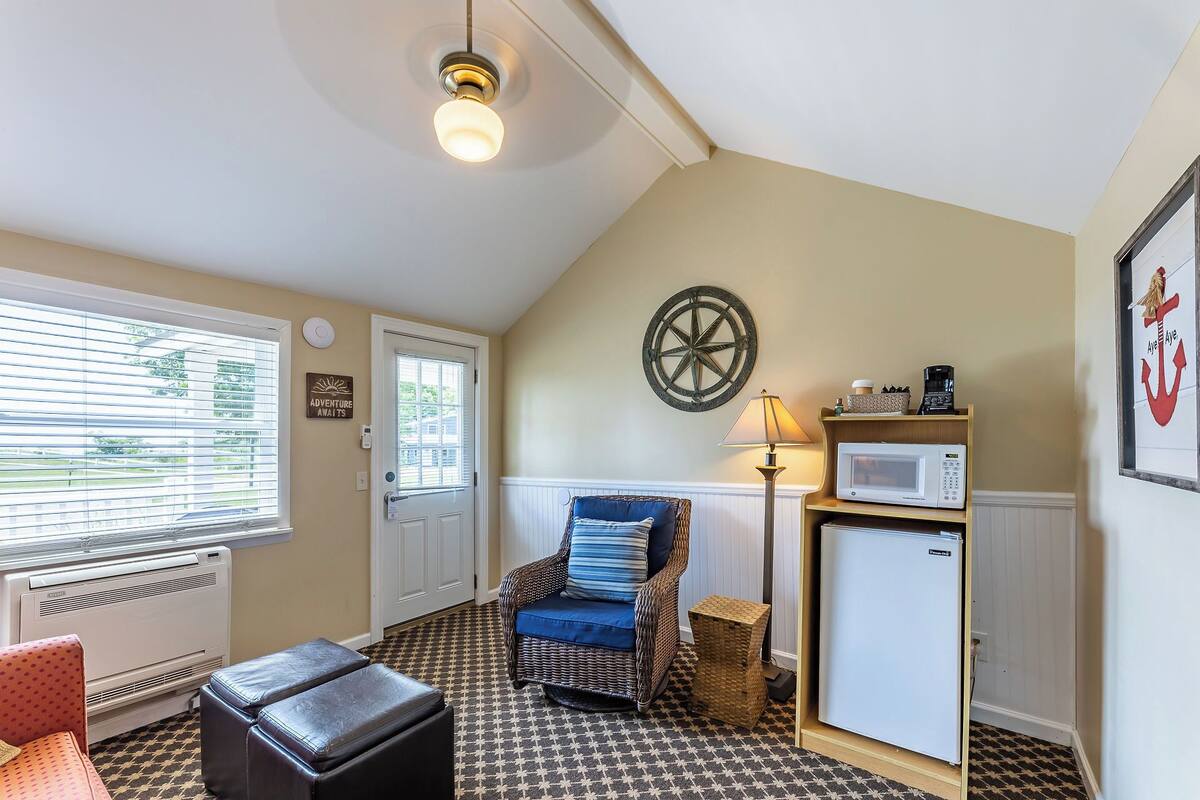A cozy room with a patterned floor, a wicker chair with blue cushions, a small white refrigerator, a wall-mounted lamp, and a door with a window.