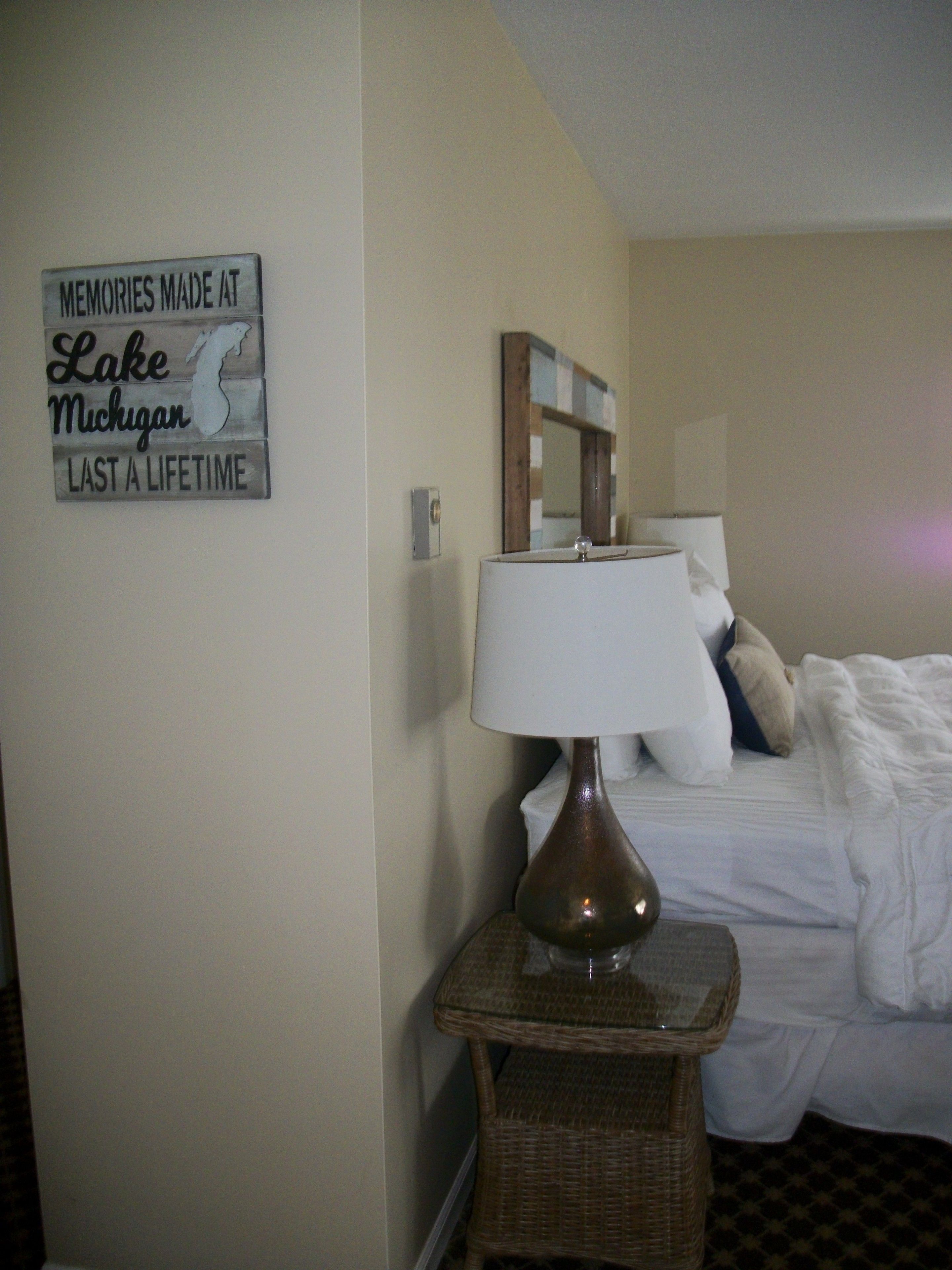 A hotel room with a bed, a bedside lamp, a small table, a mirror, and a sign on the wall.