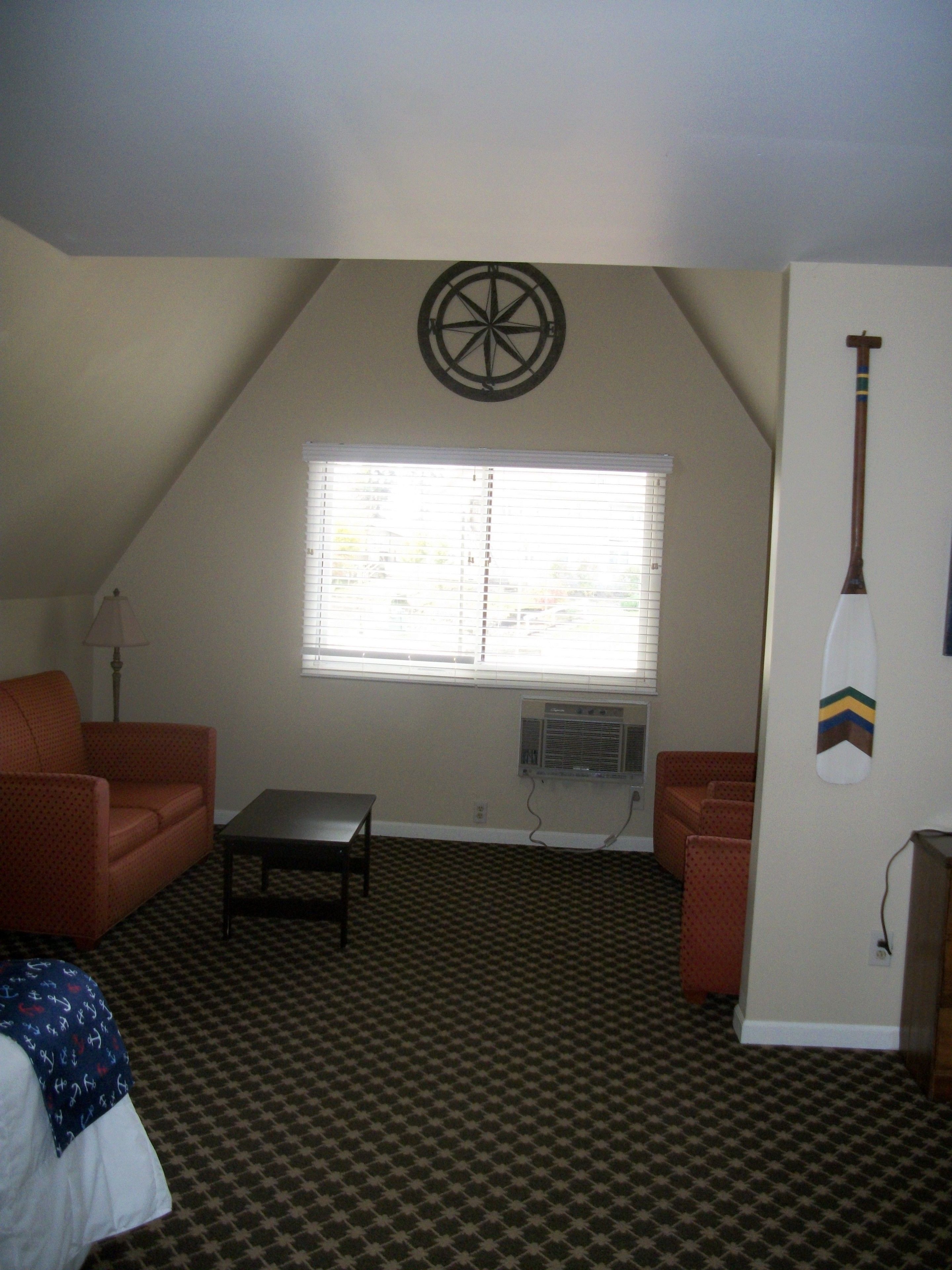 A room with a sloped ceiling, a window with blinds, a wall-mounted compass, a surfboard decoration, a patterned carpet, and two orange sofas.