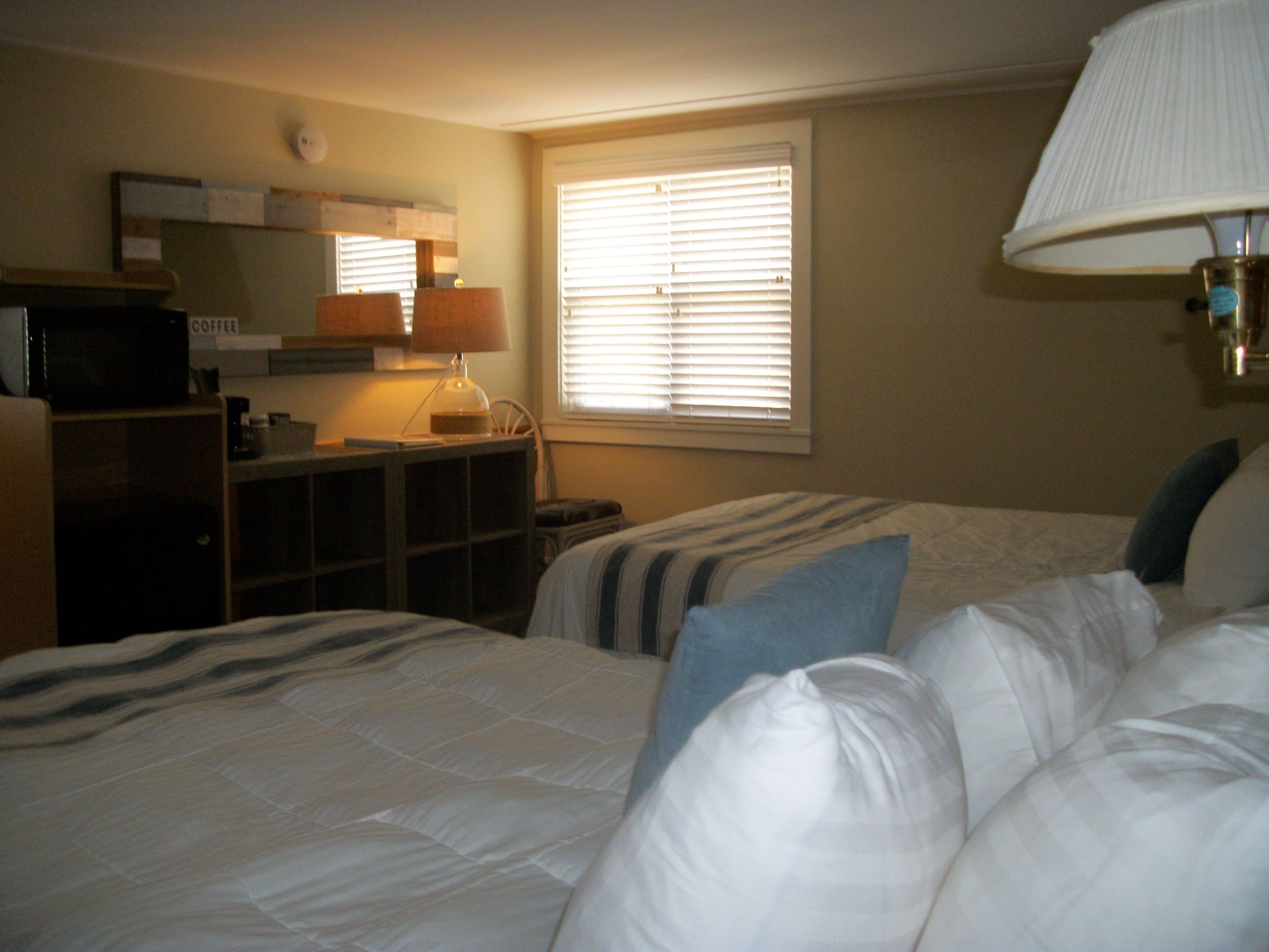Ein Hotelzimmer mit zwei Betten, einem Schreibtisch mit Lampe, einer Mikrowelle und einem Fenster mit Jalousien.