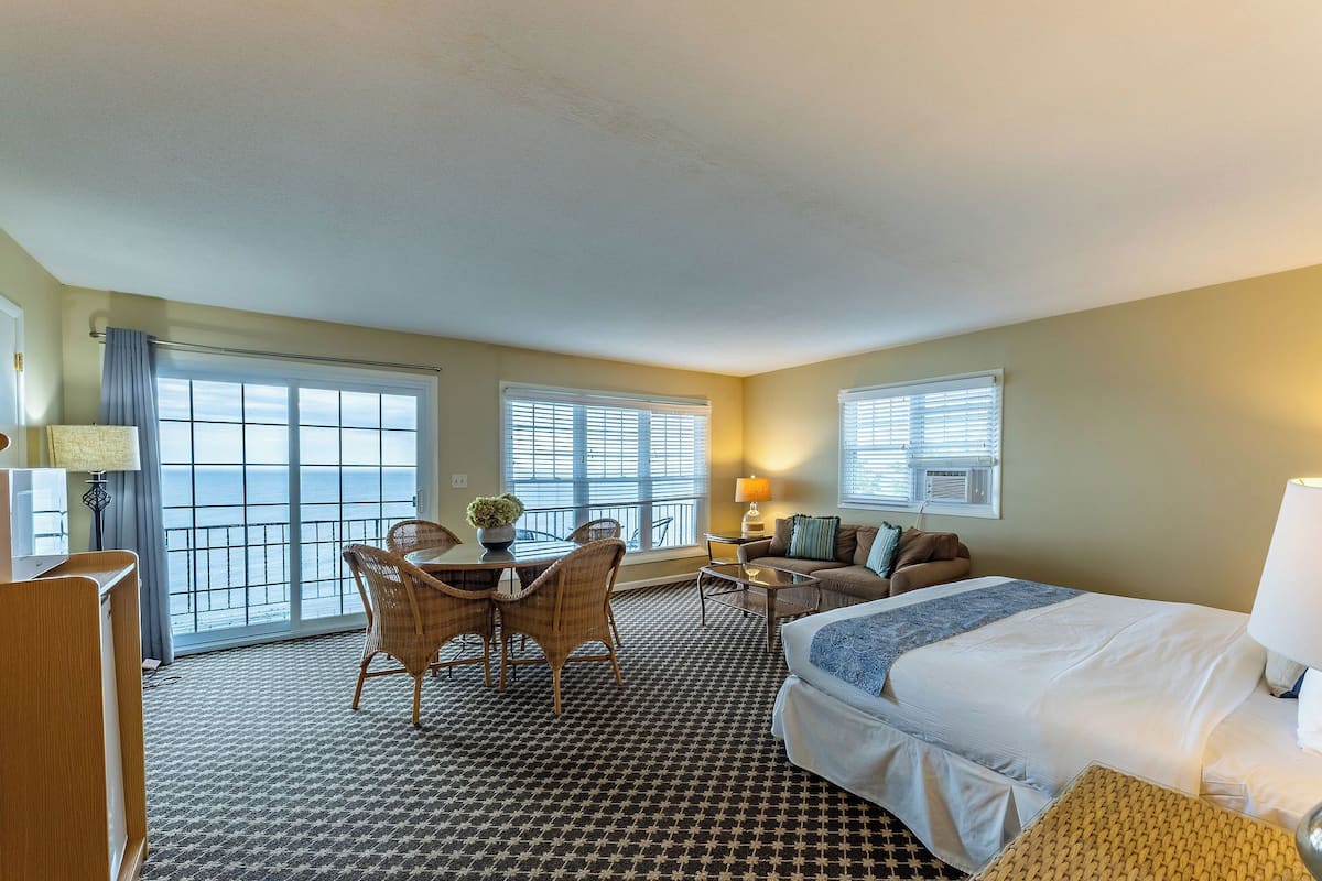 A hotel room with a bed, a sofa, a dining table with chairs, and large windows with blinds.