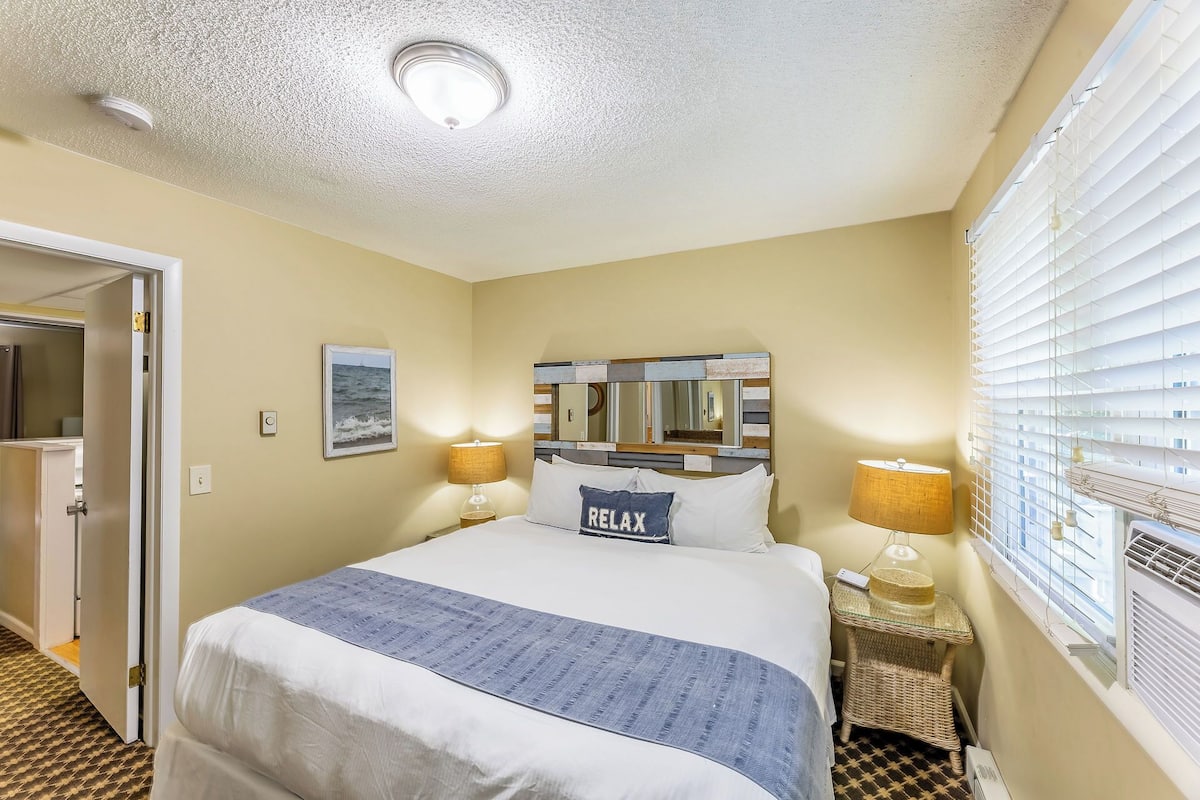 A hotel room with a large bed, two bedside lamps, a mirror with a wooden frame, a window with blinds, and a ceiling light.