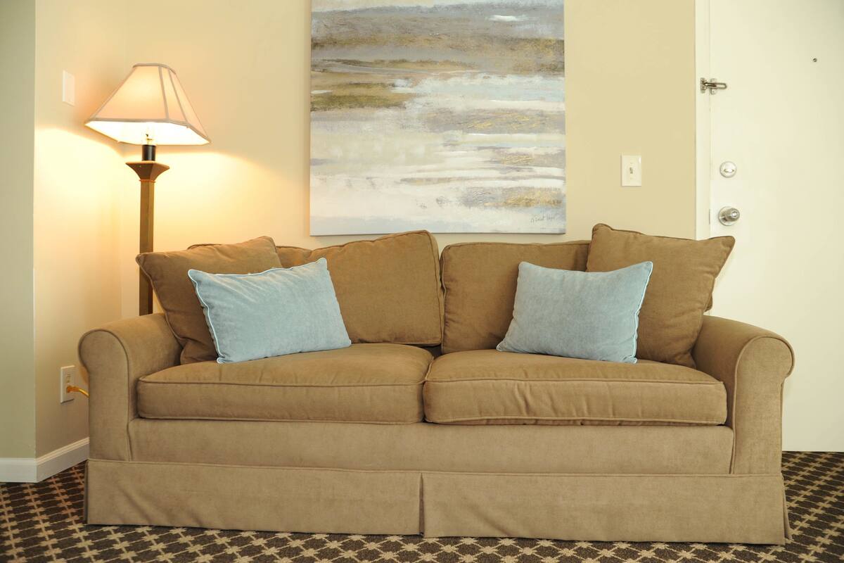 A beige sofa with two blue cushions, a floor lamp, and a painting on the wall.