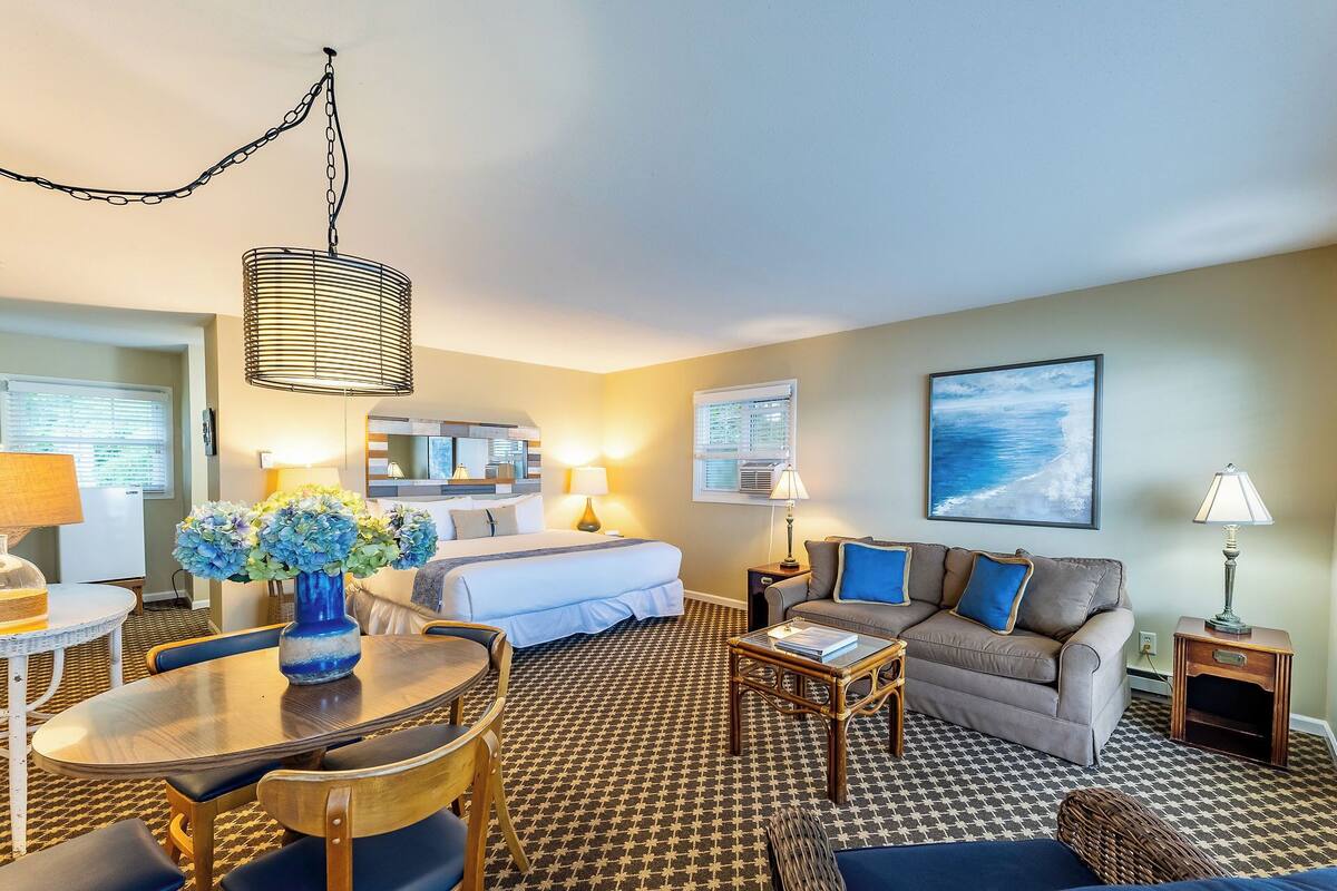 A hotel room with a bed, a sofa, a dining table, and a coffee table. There is a window with blinds, a lamp, and a painting on the wall.