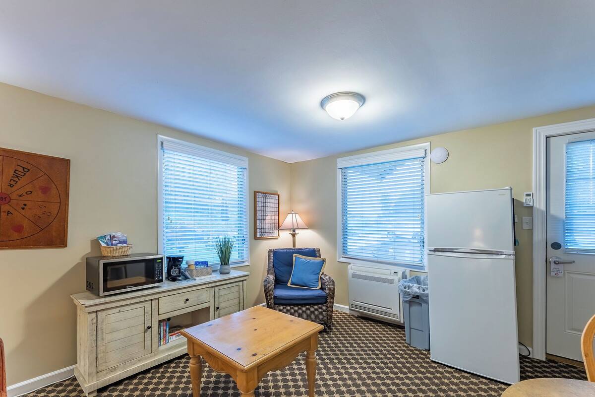 A compact kitchen with a microwave, toaster, and refrigerator. There is a dining table and a wicker chair.